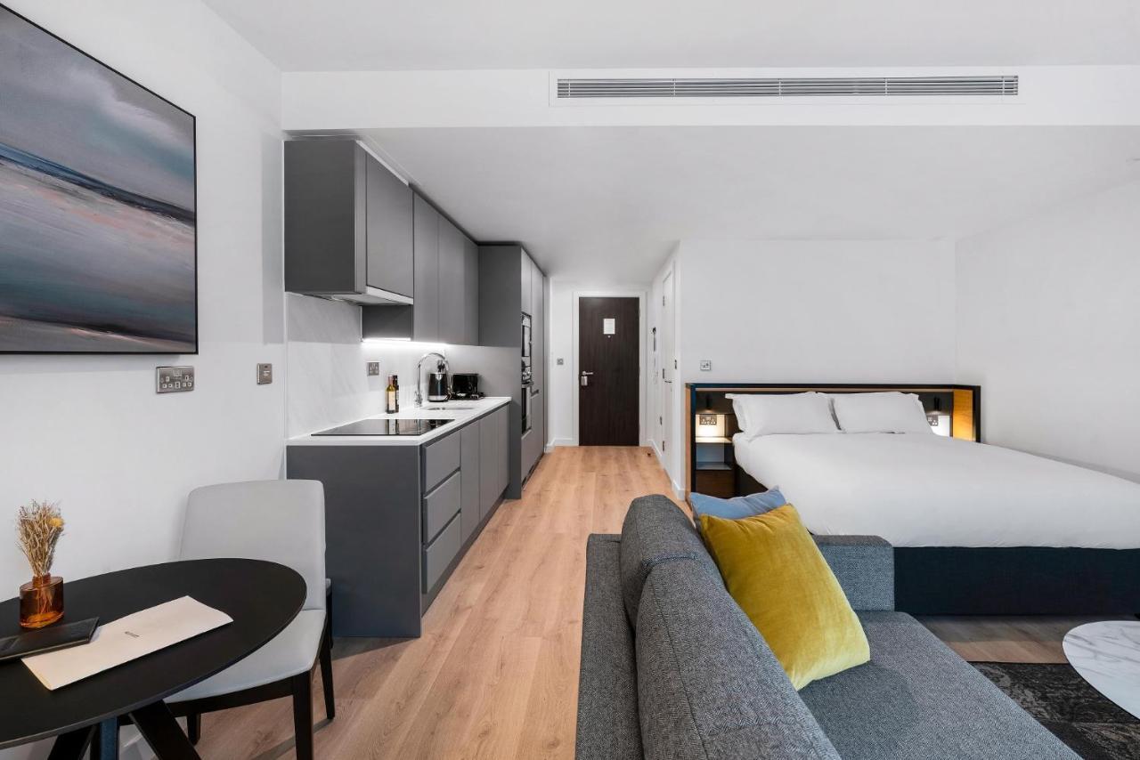 Citysuites 2 Aparthotel 맨체스터 외부 사진 The photo shows a modern and stylish studio apartment. It features a kitchenette with dark cabinetry and a small dining table with two chairs. There's a cozy sofa with colorful cushions, positioned next to the table. In the background, a large bed wi