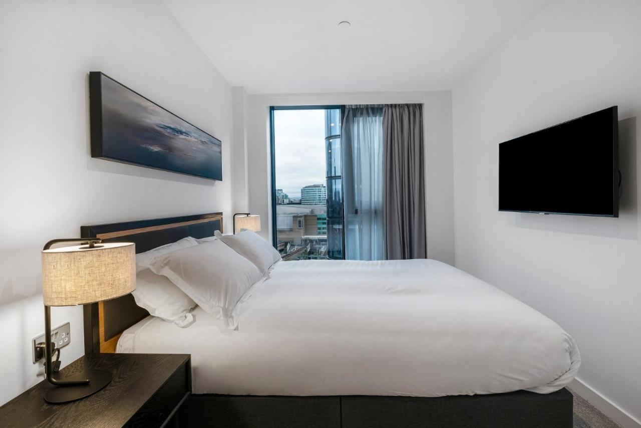 Citysuites 2 Aparthotel 맨체스터 외부 사진 The photo shows a modern bedroom with a minimalist aesthetic. It features a large bed with white bedding and two pillows. On either side of the bed, there are bedside tables with lamps. A large window lets in natural light, covered by sheer curtains.