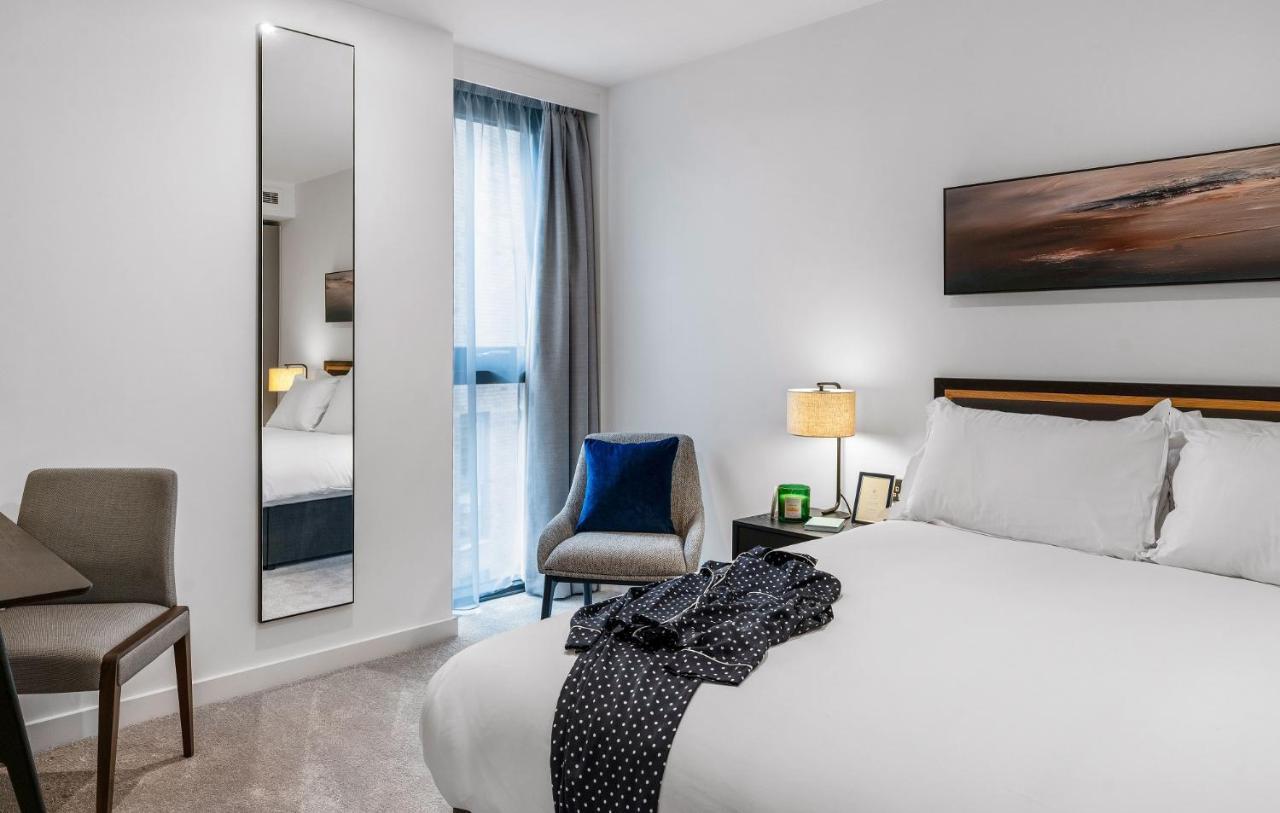 Citysuites 2 Aparthotel 맨체스터 외부 사진 The photo shows a modern hotel room. It features a neatly made bed with white linens and a dark-colored throw draped over it. There is a small bedside table with a lamp next to the bed. A comfortable chair in a light color is positioned near the wind