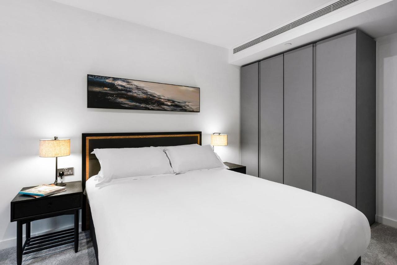 Citysuites 2 Aparthotel 맨체스터 외부 사진 The photo shows a modern bedroom designed with a minimalist aesthetic. The focal point is a large bed with white bedding, positioned against a wall. On either side of the bed, there are black nightstands topped with lamps, providing a warm glow. Abov
