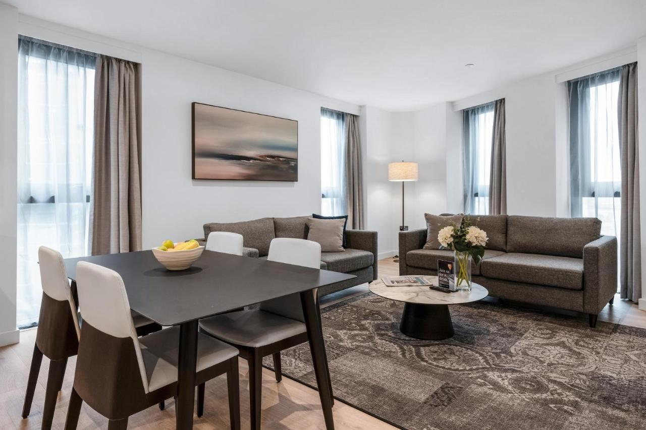 Citysuites 2 Aparthotel 맨체스터 외부 사진 The photo depicts a modern and stylish interior of a living space. On the left, there is a dining table with four chairs, featuring a bowl of fruits on the table. In the background, there's a cozy seating area with a gray sofa and a smaller round cof
