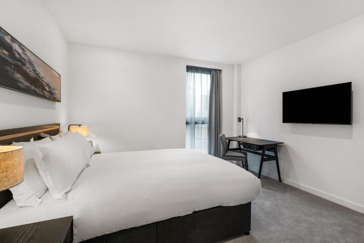 Citysuites 2 Aparthotel 맨체스터 외부 사진 The photo shows a modern hotel room featuring a large bed with white linens and a dark headboard. There are two bedside lamps on either side of the bed. A desk with a chair is placed against one wall, and a flat-screen TV is mounted on another wall. 