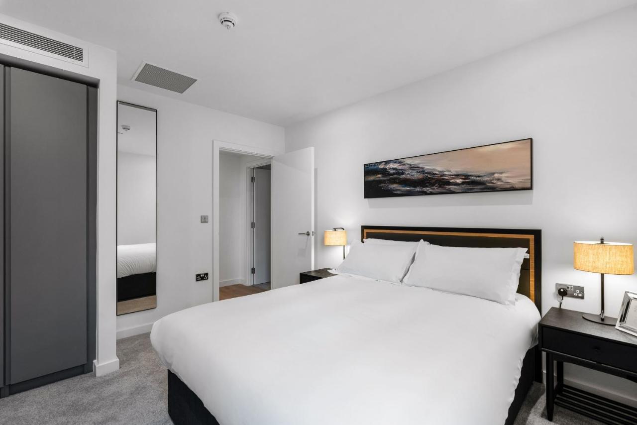 Citysuites 2 Aparthotel 맨체스터 외부 사진 The photo shows a modern bedroom featuring a large double bed with white bedding. There are two bedside tables with lamps on either side of the bed. The walls are painted white, and there's a piece of artwork above the bed that depicts a landscape. A