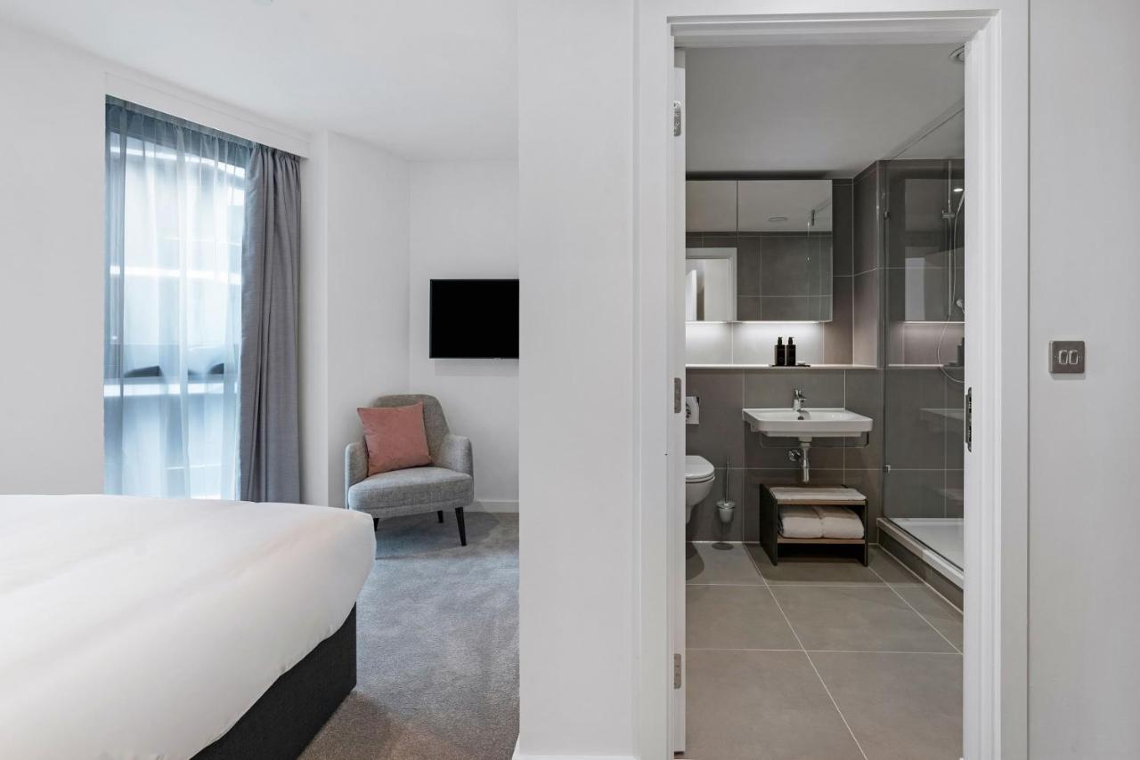 Citysuites 2 Aparthotel 맨체스터 외부 사진 The photo shows a modern hotel room and bathroom setup. On the left, there is a bed with white bedding and a small sitting area that includes a gray chair and a pillow in a subtle pink shade. A flat-screen TV is mounted on the wall, providing enterta