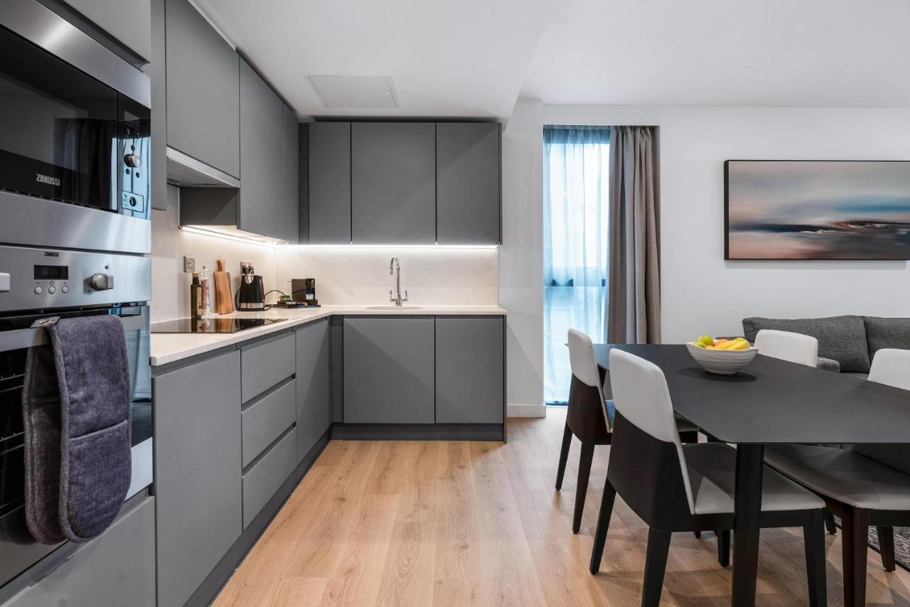 Citysuites 2 Aparthotel 맨체스터 외부 사진 The photo shows a modern kitchen and dining area. The kitchen features sleek gray cabinetry, a white countertop, and built-in appliances such as an oven. There is a sink and some kitchen utensils displayed. The adjacent dining area contains a black t