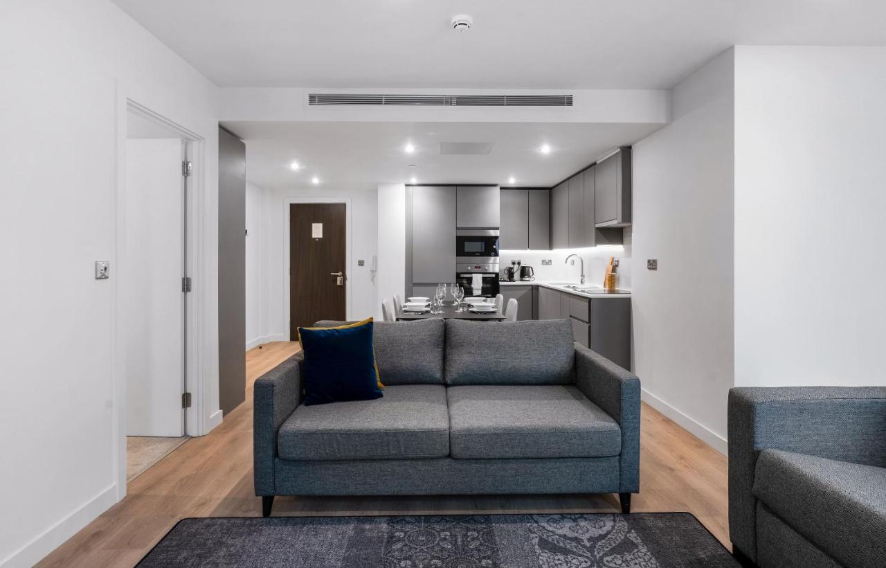 Citysuites 2 Aparthotel 맨체스터 외부 사진 The photo shows a modern interior space, likely a living area or apartment. In the foreground, there is a gray sofa with a blue cushion positioned on a textured rug. To the back, there is an open kitchen with sleek cabinetry and appliances, visible a