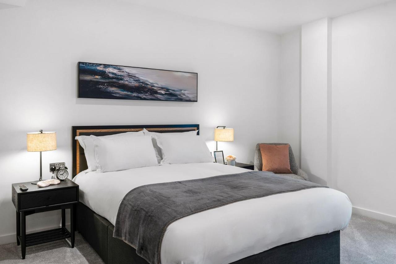 Citysuites 2 Aparthotel 맨체스터 외부 사진 The photo shows a modern bedroom featuring a large bed with crisp white linens and a grey throw blanket. On either side of the bed, there are bedside tables with lamps. A piece of abstract art hangs above the headboard, and there is a chair in a corn