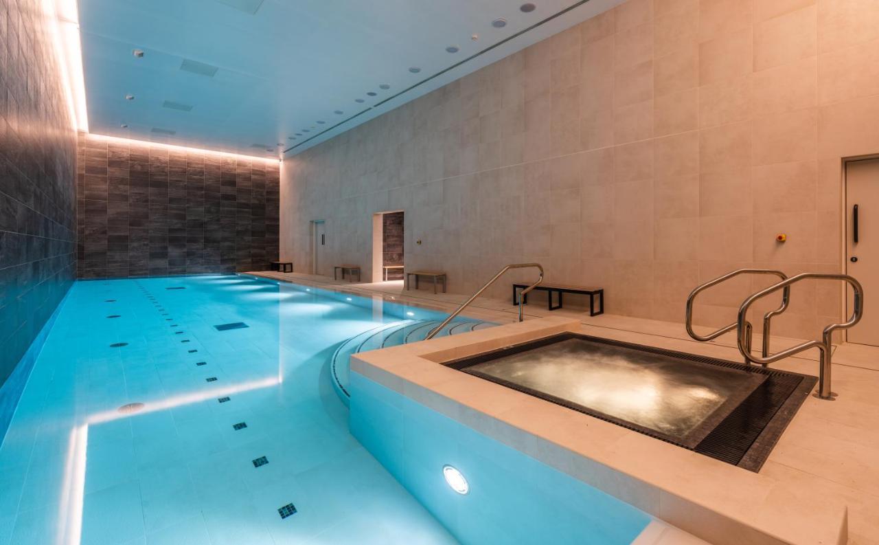 Citysuites 2 Aparthotel 맨체스터 외부 사진 The photo shows an indoor swimming pool area. The pool is a bright blue color and features smooth, tiled surfaces. On one side, there is a hot tub area with steps leading into it. The walls are made of light-colored stone, and there is ambient lighti
