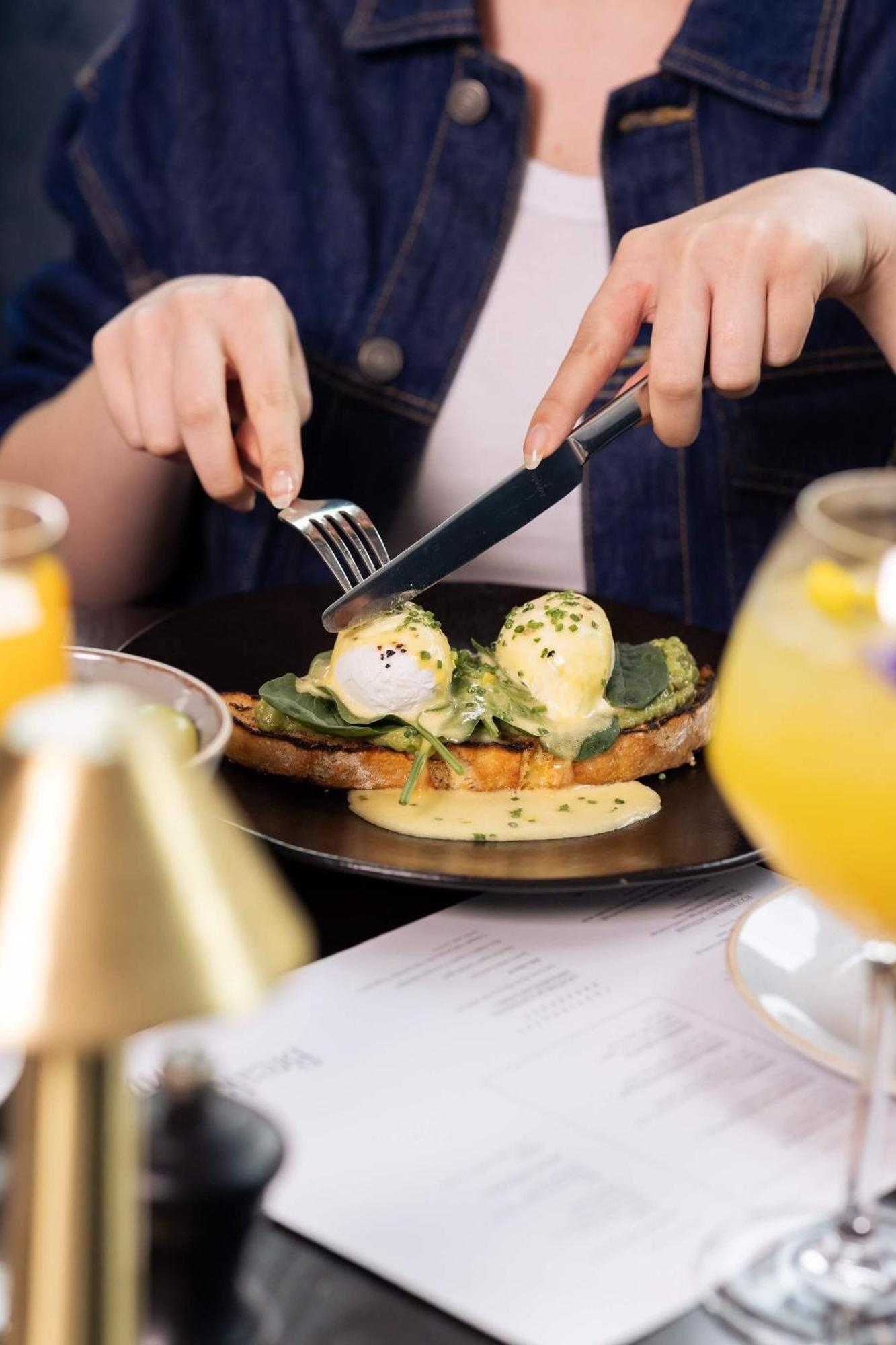 Citysuites 2 Aparthotel 맨체스터 외부 사진 The photo shows a person preparing to eat a dish of eggs Benedict. They are holding a knife and fork, ready to cut into the meal, which features poached eggs on top of a slice of toast, accompanied by spinach and a creamy hollandaise sauce drizzled o